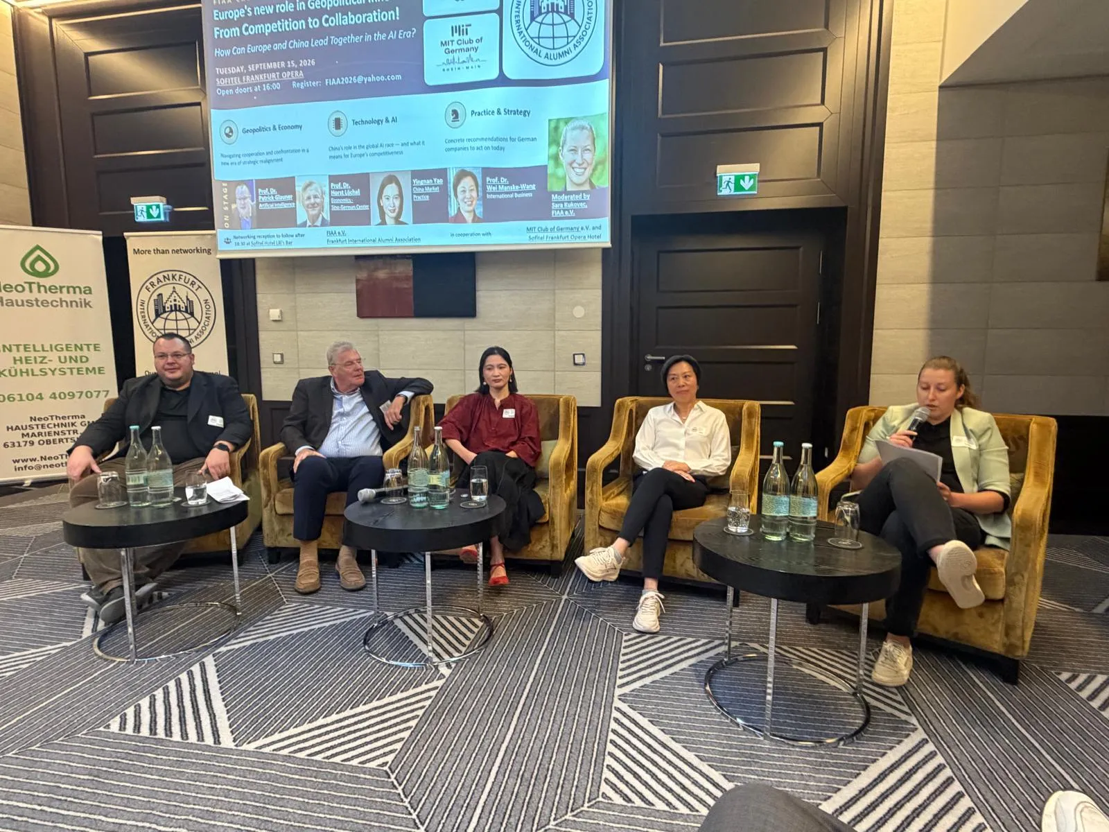 Das Panel auf der Bühne im Sofitel Frankfurt Opera, im Hintergrund die Veranstaltungsfolie mit den vier Panelist:innen und der Moderation
